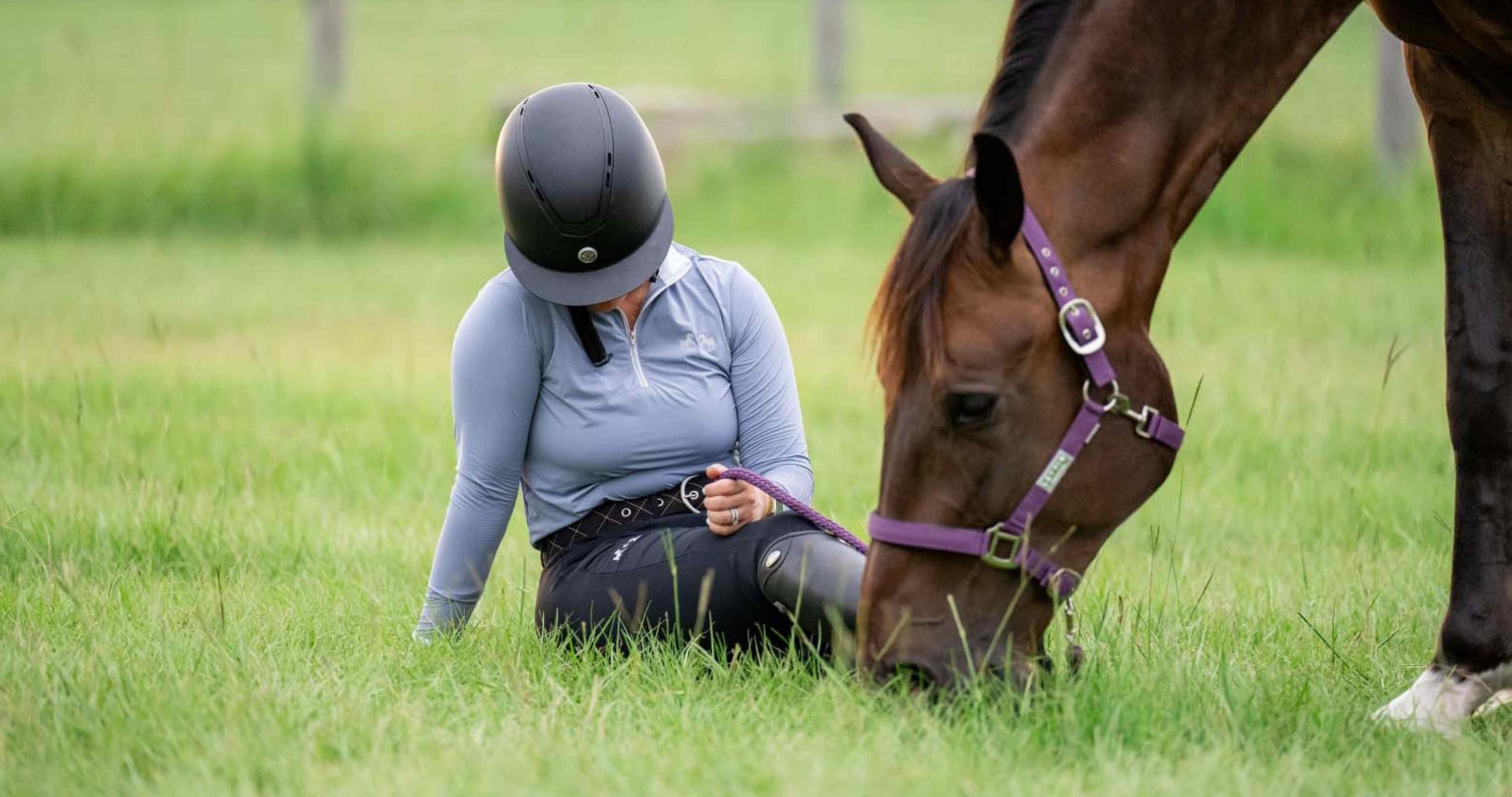 sun shirt, UV protection clothing, best UV protection for horses, female rider wearing black wide brim helmet and sun shirt lounges in grass while handgrazing bay horse wearing purple halter