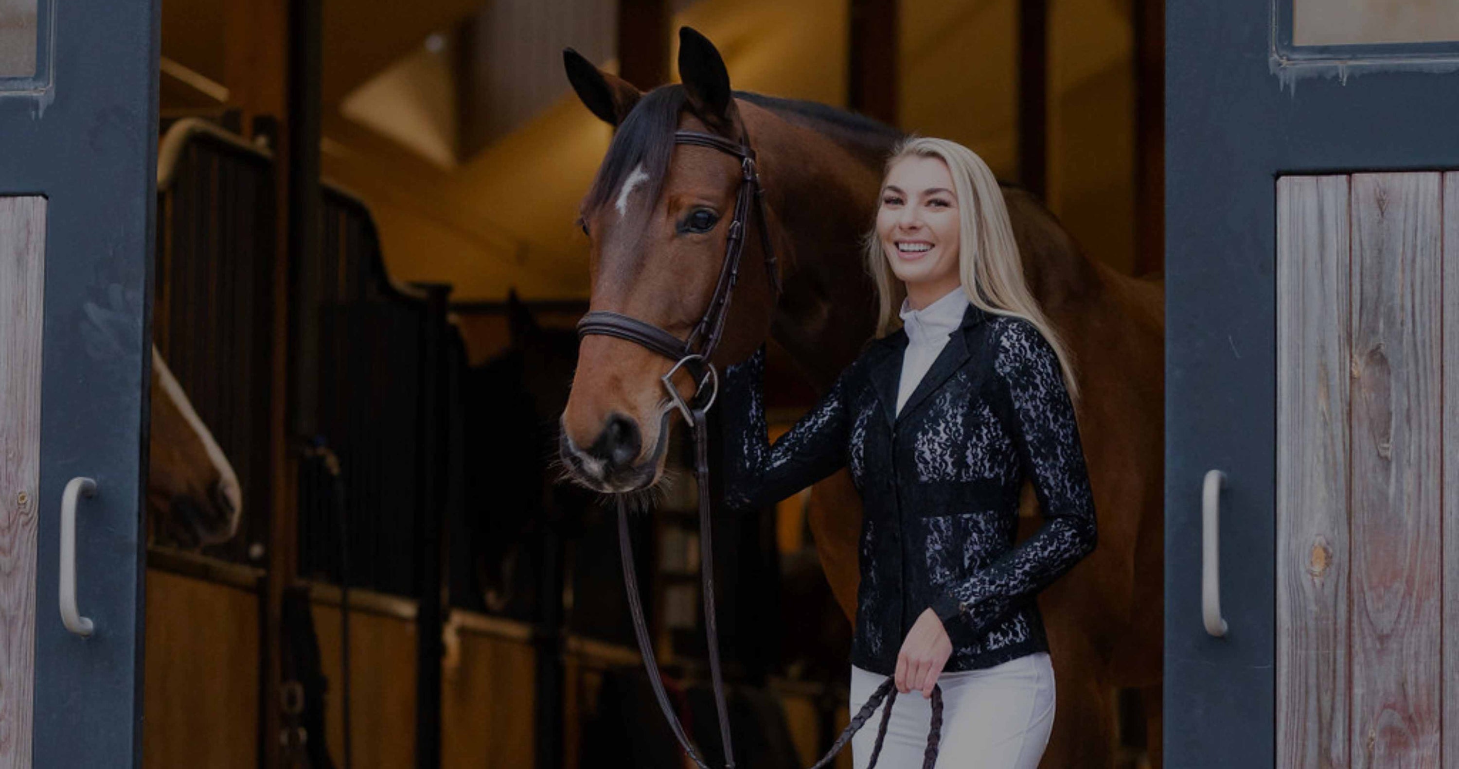 English horse show clothes, blonde woman wearing English horse clothes stands with bay horse in barn aisle. 