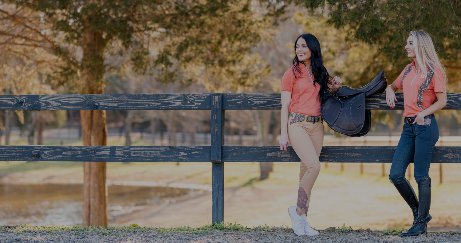 A blonde and brunette woman wearing EcoRider clothing in pink and navy stand by a wooden fence with a saddle.