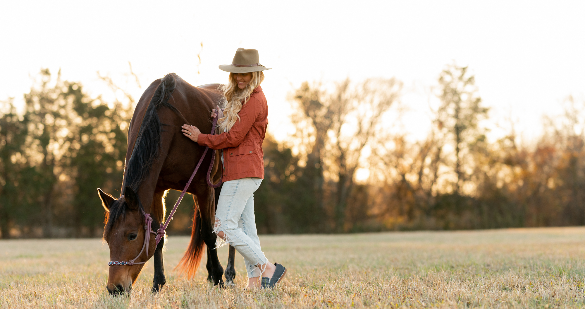 western style woman wearing orange shirt and blue jeans stands next to bay horse during the golden hour